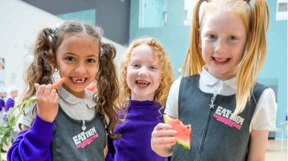 Three young school girls wearing Eat Them to Defeat Them badges smile as they hold up fruits and vegetables.