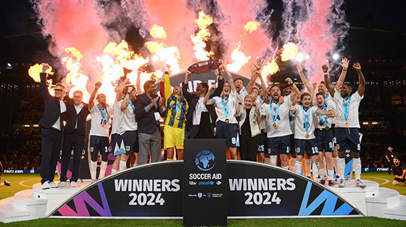 Soccer Aid football players celebrate on the pitch. They are standing on a stage that says “Winners 2024” in front of flame cannons. They hold up the trophy smiling and cheering with their arms.