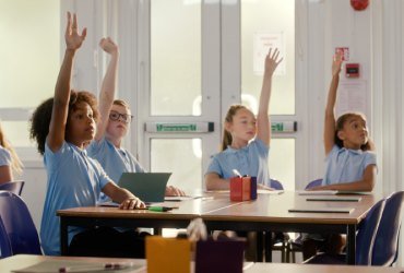 Four school-children sit in a classroom with their hands raised as if to answer a question set by the teacher.