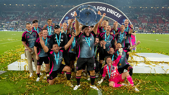 Soccer Aid football players celebrate on the pitch at Old Trafford. They are standing in front of a stage with a banner behind them that reads “Soccer Aid for Unicef”. At the front of the football squad is Big Zuu holding up the winning silver plate.