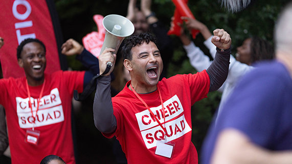 Actor Ryan Thomas cheering on people at a sporting event. He has both hands in the air and is carrying a megaphone in his right hand. He is wearing a t-shirt that reads “Cheer Squad” and a lanyard. Behind him is another man wearing the same outfit who is also cheering.