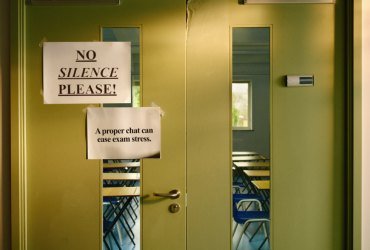 A 'No Silence Please' sign is attached to a set of double doors that have windows. Another sign reads 'Supported by Mind, YoungMinds and ITV.' Through the windows, there are rows of desks and chairs set up in an exam room style.